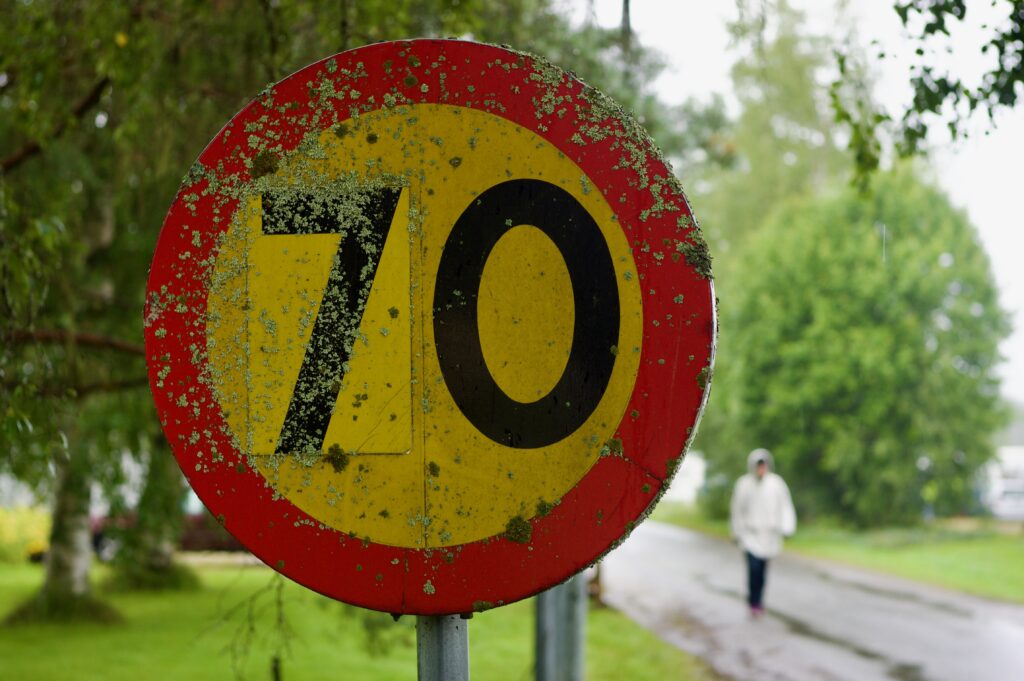 I front, et svenskt 70 km/t hastighetsskilt, hvor tallet 7 er klistret over et annet tall. Skiltet har rød sirkel og gul bakgrunn. Det gror mose på skiltet. Uskarpt i bakgrunnen går det en person i hvis jakke i regnet.